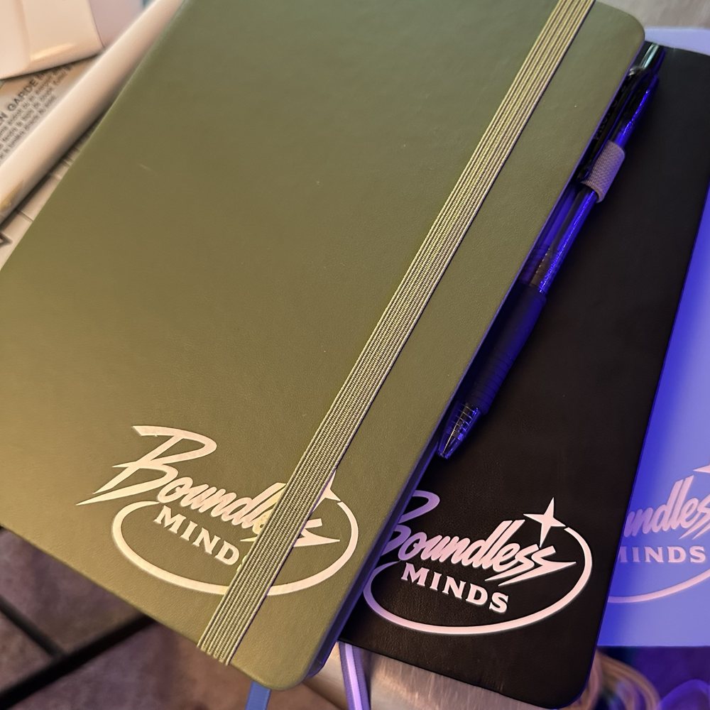 Notebooks with the Boundless Minds logo in white: an olive green one with an elastic band on top of a black one with a pen in its loop